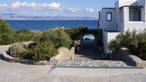 A white building with a stone archway leading to a patio with a table and chairs, overlooking a body of water and mountains. -  Villa With Swimming Pool In Front Of Almost Private Beach  (PAROS)