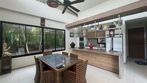 A modern kitchen with a central island, a dining area with wicker chairs, and a large window with a view of greenery.