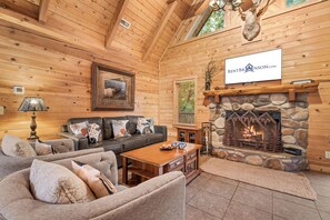 L’intérieur d’une cabane en rondins, avec une cheminée en pierre, des canapés en cuir et un tableau représentant un cerf, encadré.