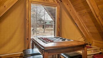 A cozy attic room with a wooden sloped ceiling, a large window with blinds, a wooden table with a board game, and two leather benches.