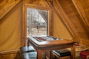 Een gezellige zolderkamer met een houten schuine daklijn, een groot raam met zonwering, een houten tafel met een spel, en twee leren bankjes.