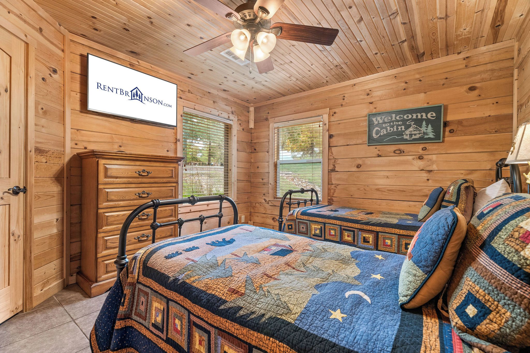 A cabin interior with wooden walls, a bed with a quilt, a dresser, and a sign that reads 'Welcome to the Cabin.'