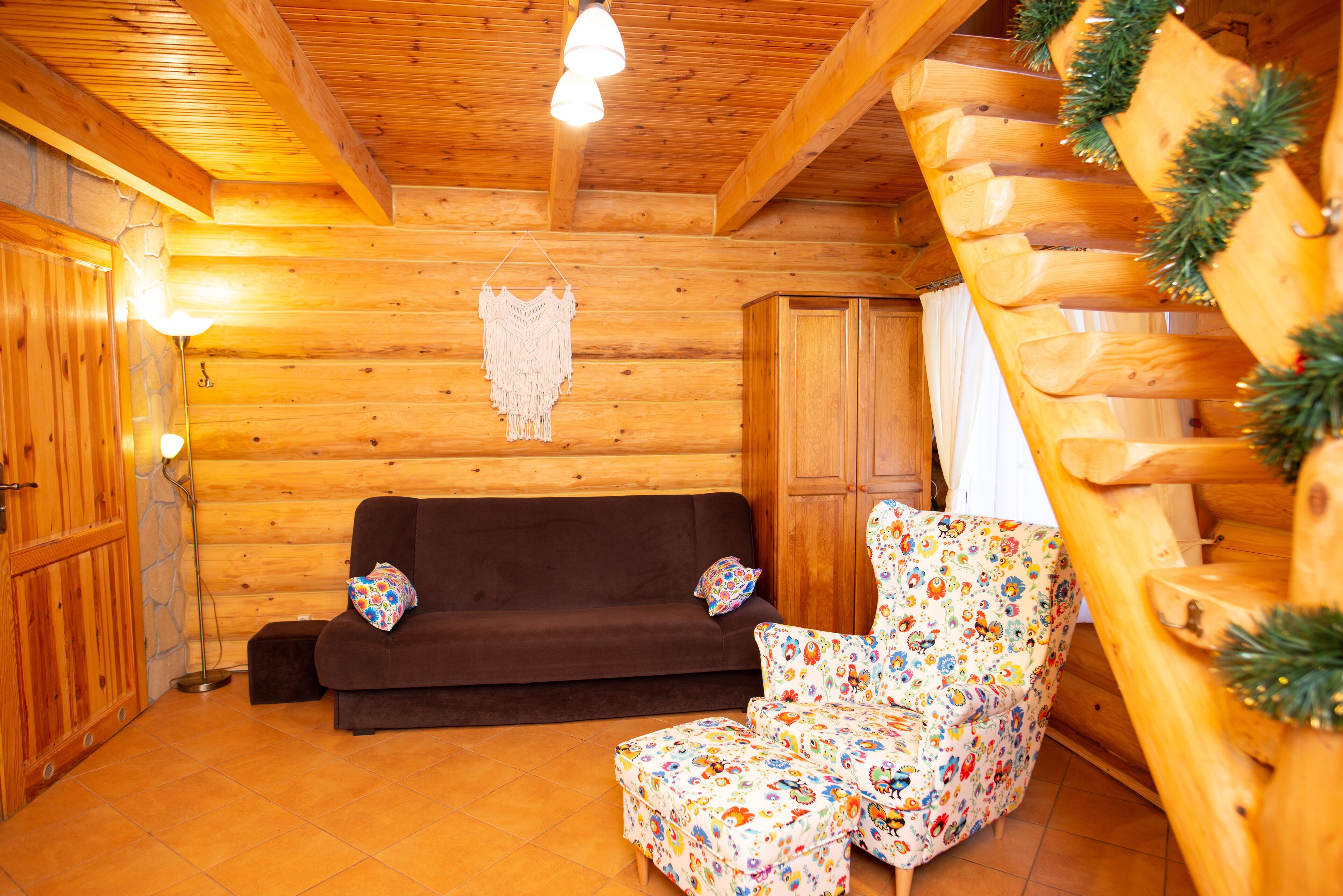 Un interior acogedor de cabaña de madera con un sofá marrón oscuro, un sillón estampado con flores y un tapiz decorativo de macramé.