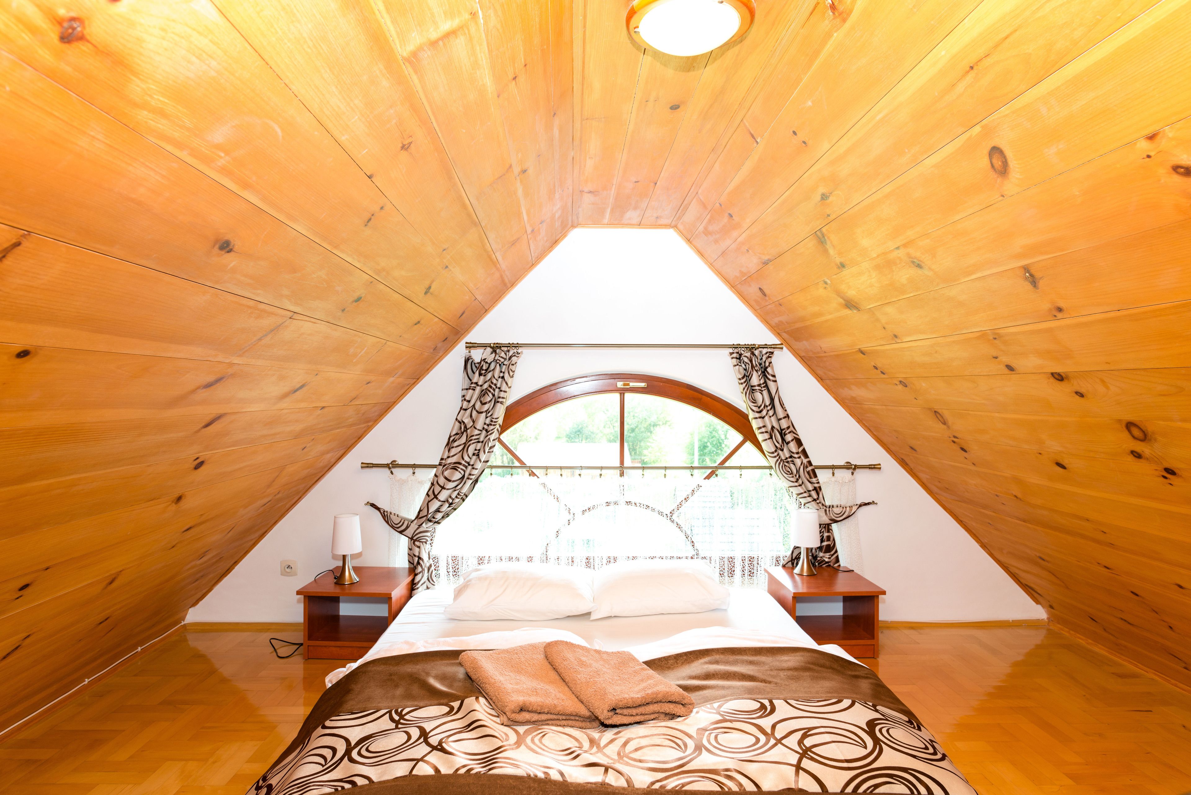 A bedroom with a sloped ceiling, a bed with a patterned bedspread, a nightstand with a lamp, and a window with curtains.