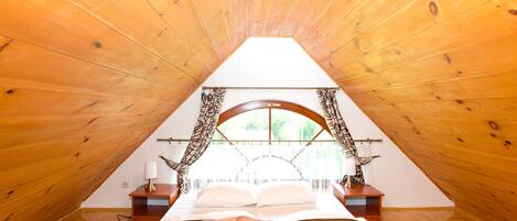 A bedroom with a sloped ceiling, a bed with a patterned bedspread, a nightstand with a lamp, and a window with curtains.