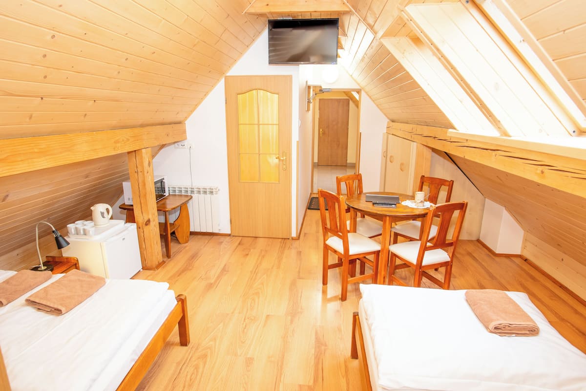 Ein gemütliches Schlafzimmer im Dachgeschoss mit Holzdielen, einem Esstisch mit Stühlen, einer kleinen Küchenzeile und einem an der schrägen Decke befestigten Fernseher.