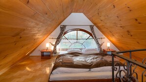 Un dormitorio abrigado en el ático, con techo inclinado de madera, una cama grande con colcha estampada, dos lámparas de noche y una ventana con cortinas.