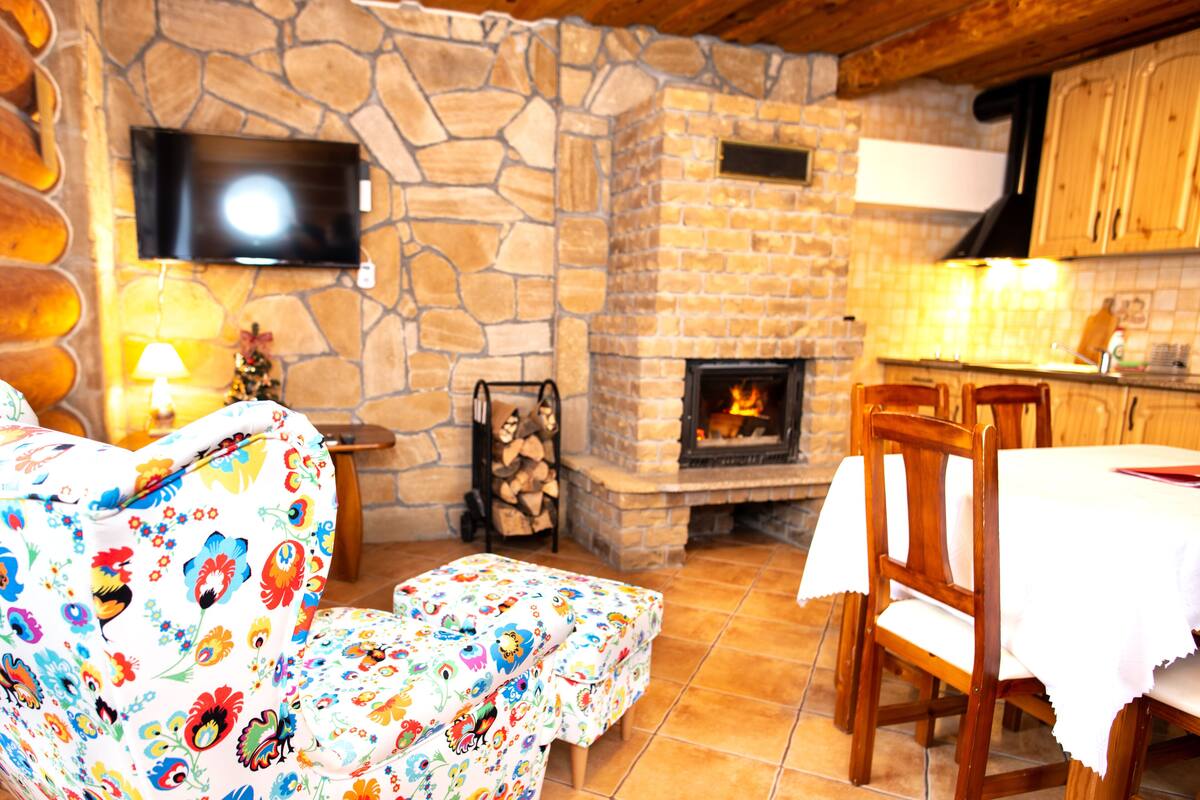 Ein gemütliches Wohnzimmer mit Kamin aus Stein, einem an der Wand befestigten Fernseher und einer Essecke mit Holztischen und -stühlen.