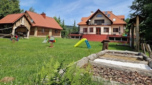 Una casa grande con techo rojo, una cabaña de madera y un patio con césped y juegos infantiles.