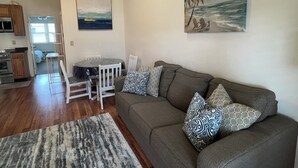 A living room with a gray sofa, patterned pillows, a dining table with chairs, a painting of a beach scene, and a kitchen area with wooden cabinets and a ceiling fan.