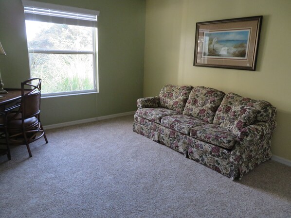 A room with a floral-patterned sofa, a wooden chair, a desk, and a window with blinds. - Beautiful 3/2 designer home, pet friendly, free WiFi, golf cart, hot tub (The Villages)