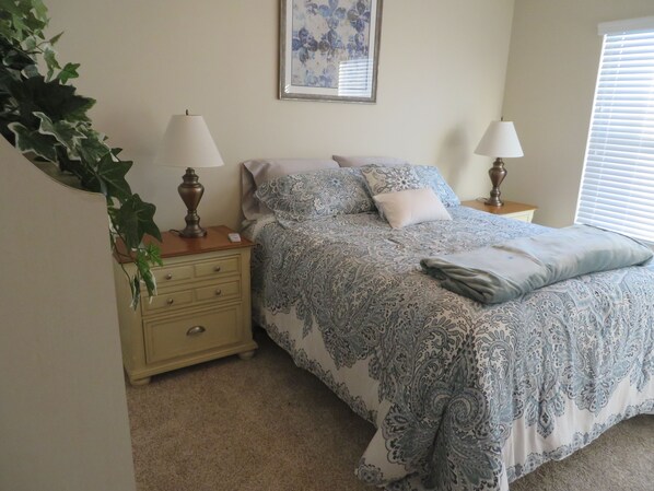 A bedroom with a bed, two bedside tables, a nightstand, a wall-mounted painting, and a window with blinds. - Beautiful 3/2 designer home, pet friendly, free WiFi, golf cart, hot tub (The Villages)
