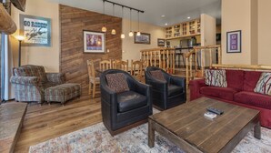 A cozy living room with a wooden floor, a red sofa, black leather chairs, and a coffee table. There are wooden chairs and a dining table in the background. - Short Walk to Slopes, Private W/D + Pool & Hot Tub (Steamboat Springs)