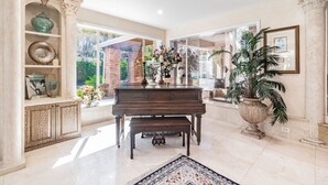 Um piano de cauda imponente em uma sala ampla, com grandes janelas, um aparador decorativo e um vaso com planta.