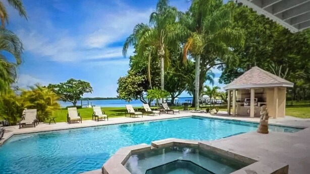 Piscina com espreguiçadeiras e ofurô, cercada por palmeiras e um gazebo.