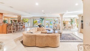 Uma sala de estar espaçosa com um amplo sofá modular bege, uma área de jantar com balcão e vista para uma piscina e vegetação através de grandes janelas.