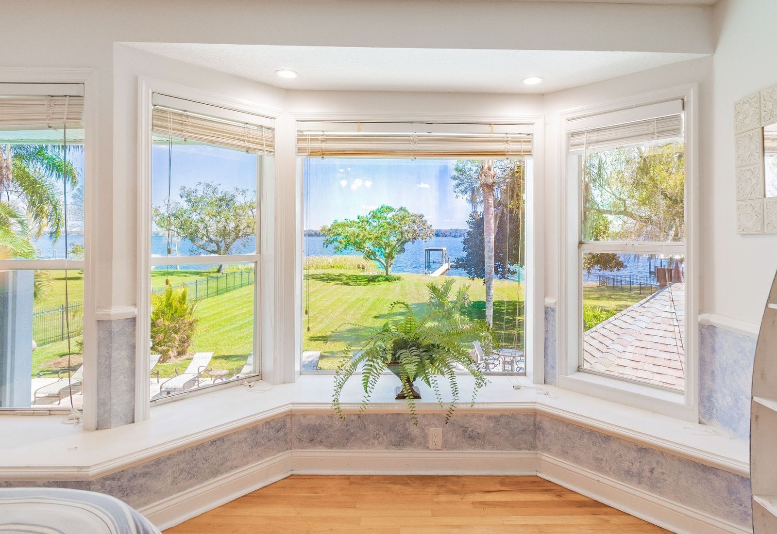 A room with a bay window overlooking a landscaped garden and a body of water.