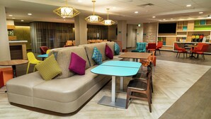 A modern hotel lobby with a seating area featuring a sofa and tables, a bar area with a counter and seating, and a large screen on the wall. - Home2 Suites by Hilton Roseville Sacramento (Roseville)