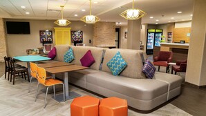 A modern hotel lobby with a sofa, orange ottomans, a table with chairs, and a reception desk. - Home2 Suites by Hilton Roseville Sacramento (Roseville)