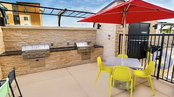 An outdoor patio with a stone wall, a grill, yellow chairs, and a red umbrella.