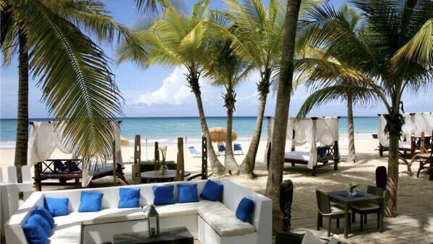 Een strandlounge met een witte bank, blauwe kussens en een houten tafel.