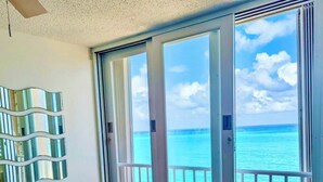 Een woonkamer met een donkergrijze bank, een kleine witte tafel en een potplant. Een groot schuifglas raam leidt naar een balkon met uitzicht op zee en strand.