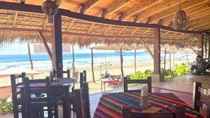 An outdoor seating area with wooden tables and chairs, a thatched roof, and a view of the beach and ocean. - Posada Mexico (San Pedro Pochutla)