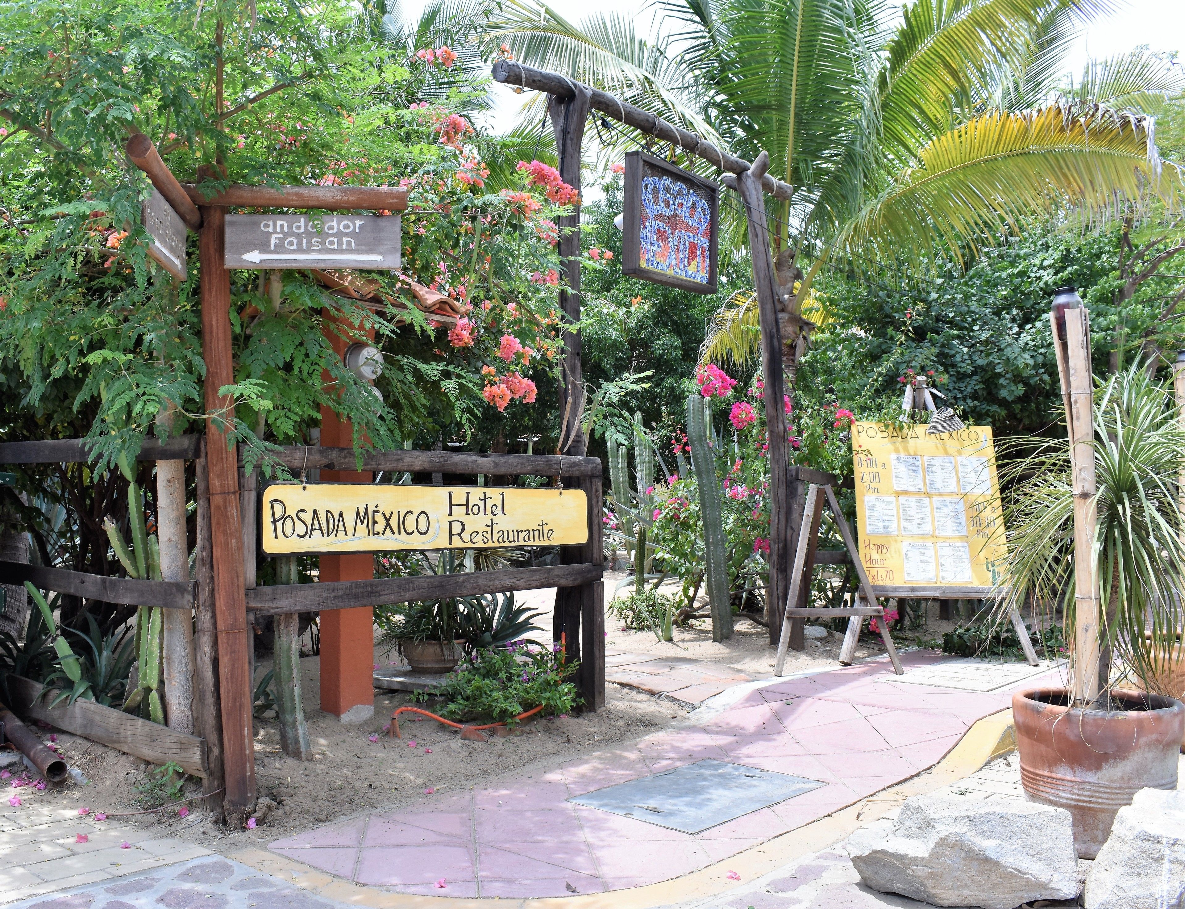 Un cartello in legno con la scritta "Posada Mexico Hotel Restaurante" e "Andador Faizan", circondato da vegetazione lussureggiante e cactus.