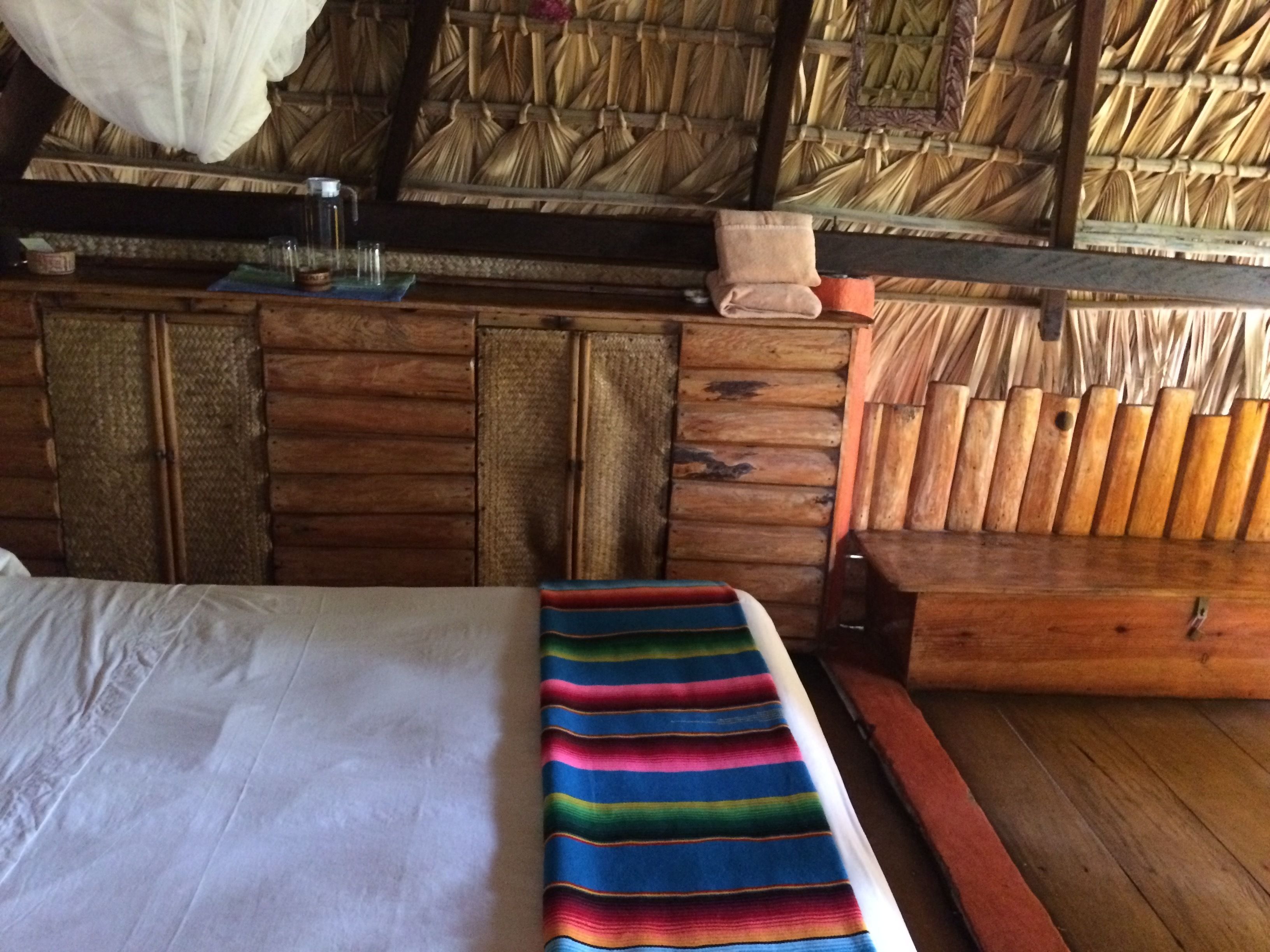 A room with a bed, a colorful striped bench, wooden furniture, and a woven wall.
