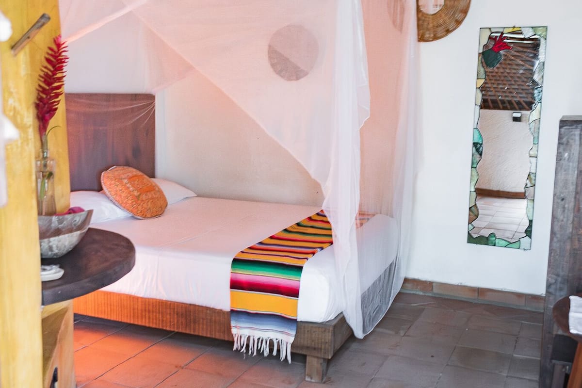 A room with a bed under a canopy, a wooden headboard, a small table with a vase, and a mirror on the wall.