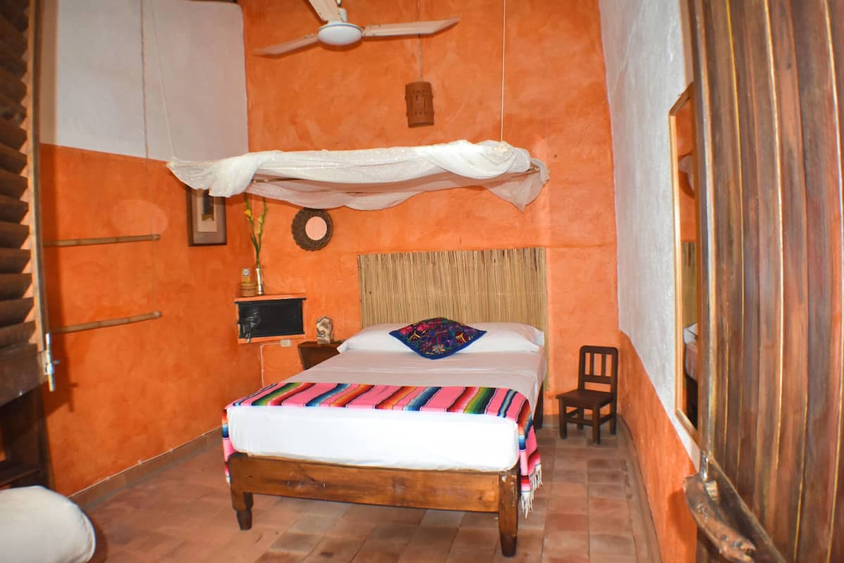 A room with an orange wall, a wooden bed with a colorful blanket, a small bedside table with a lamp, a chair, and a ceiling fan.