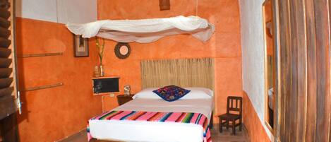 A room with an orange wall, a wooden bed with a colorful blanket, a small bedside table with a lamp, a chair, and a ceiling fan.