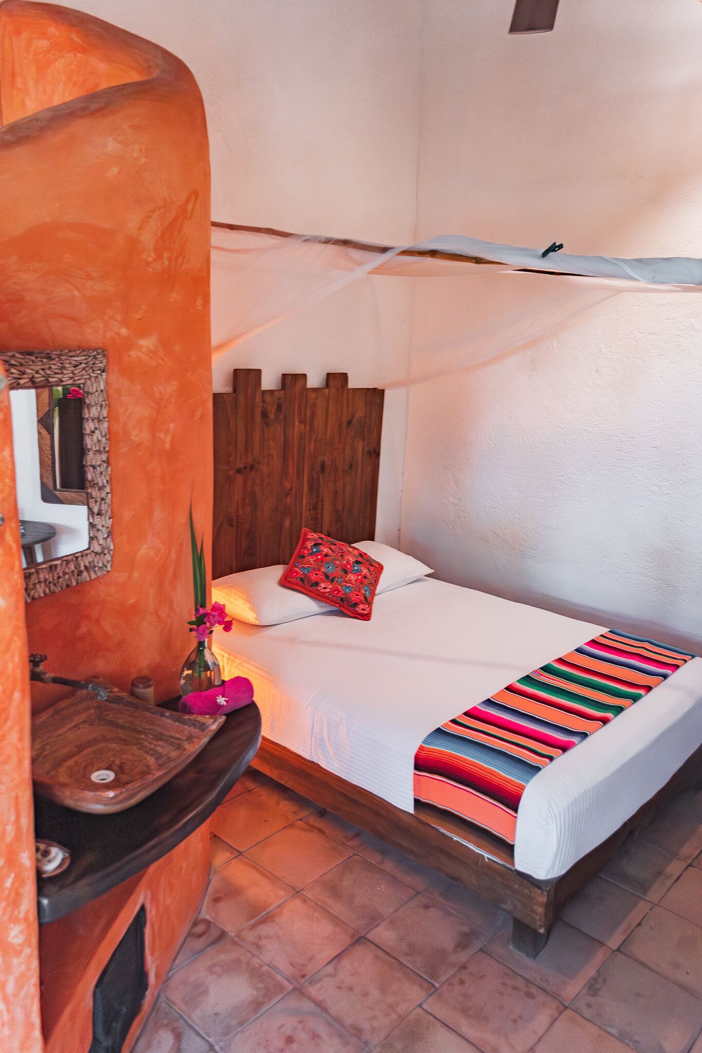 A small, cozy room with a bed, a wooden headboard, a bedside table with a mirror, and a colorful striped blanket.