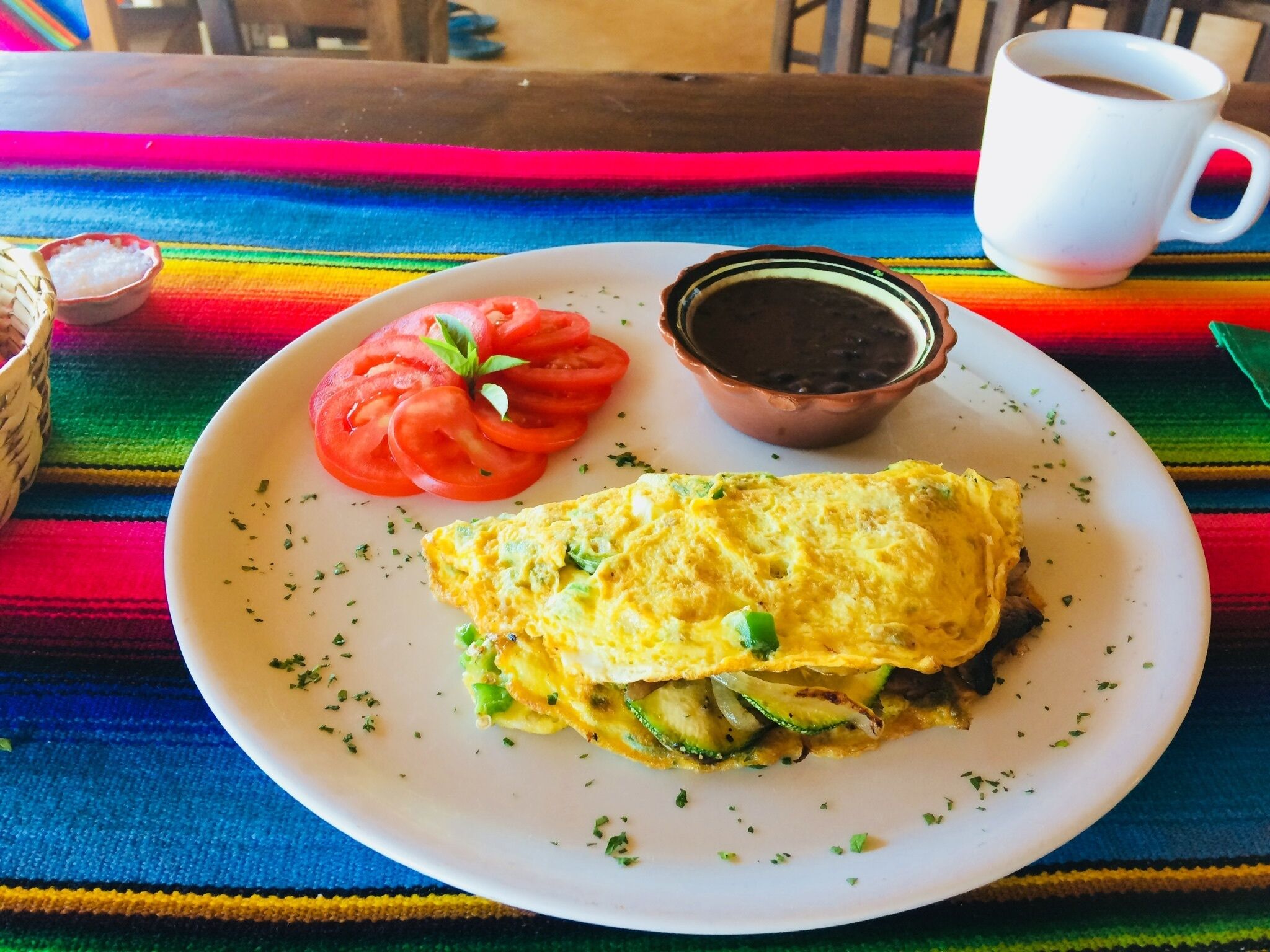 Une assiette contenant une omelette, des tomates en tranches et un bol de haricots noirs, le tout posé sur une nappe aux couleurs vives.