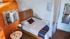 A bedroom with a wooden headboard, a white bed with a colorful striped blanket, a white canopy, a wooden bedside table with a bowl and a flower, and a mirror with a decorative frame. - Posada Mexico (San Pedro Pochutla)