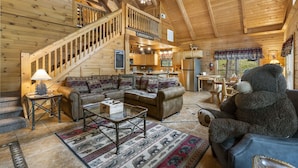 A cozy cabin interior with a wooden staircase, a living room with a sofa and a large teddy bear, a kitchen area, and a dining table. - Secluded Lodge w HotTub_Loft w Games_Near Big Cedar Trophy Buck_Pet Friendly_Car Charger_Slide (Ridgedale)
