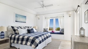 Une chambre à coucher comprenant un lit, des tables de chevet, une fenêtre avec des rideaux, un ventilateur de plafond et une vue sur l’extérieur.