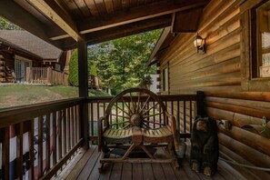 A wooden porch with a rocking chair, a small bear statue, and a window with a view of a house and trees. - Upscale Log Cabin Over 2400 Sq Ft- Game Table-Free Tickets-Hot Tub-Car Charger-Near Thunder Ridge (Ridgedale)