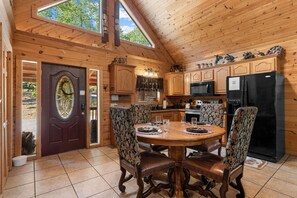 A wooden cabin interior with a dining table, chairs, kitchen appliances, and a large window.