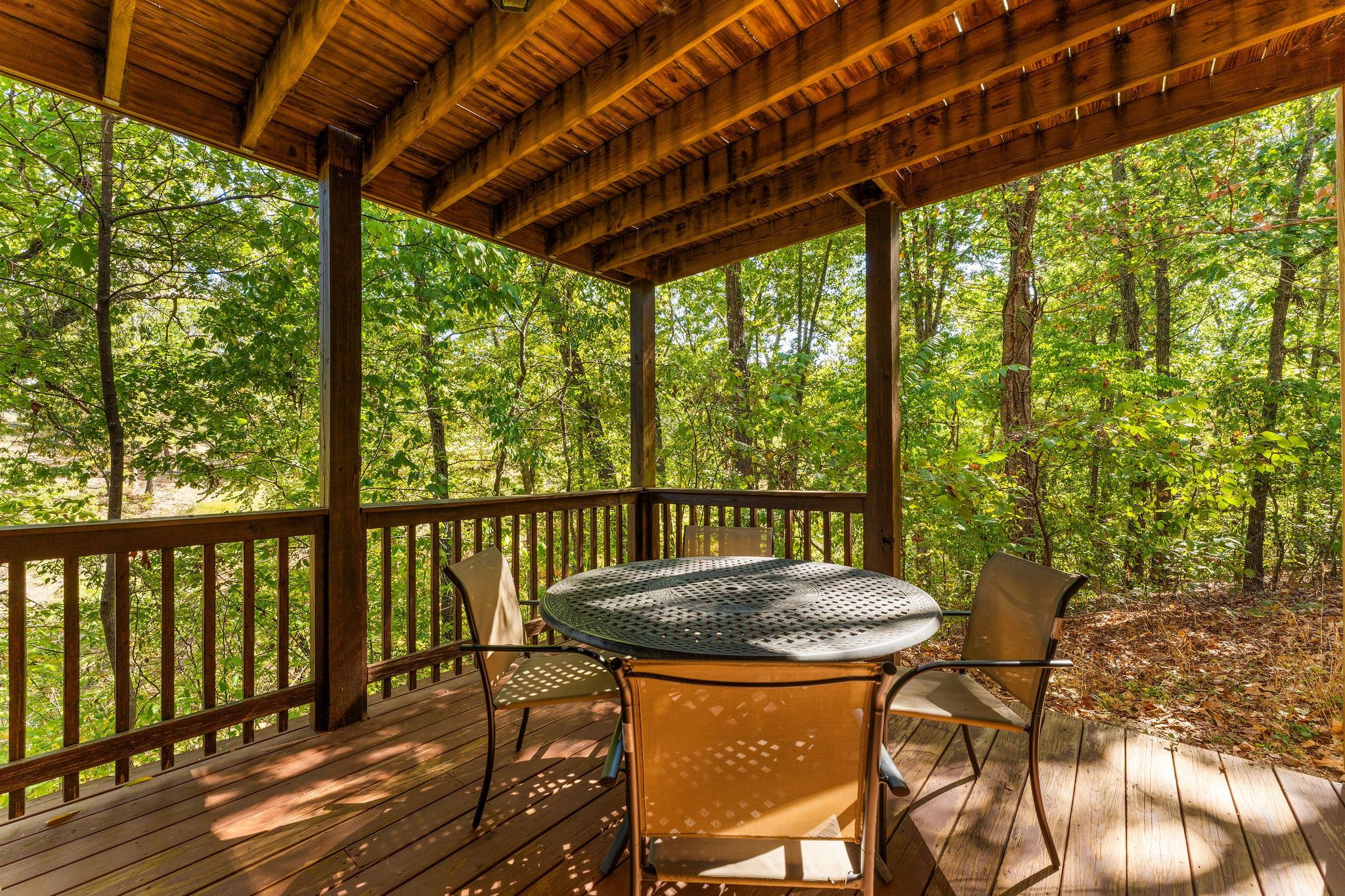 A wooden deck with a round table and two chairs, surrounded by a wooden railing, set in a forested area.