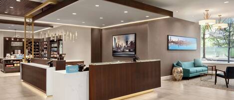 A modern hotel lobby with a reception desk, seating area, and a large window providing a view of the outdoors.