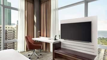 A hotel room with a large window, a flat-screen TV mounted on the wall, a desk with a chair, and a bed.