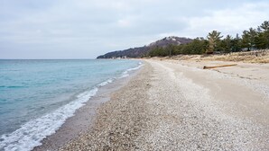 A pebbly beach with calm waters, a wooden log on the sand, and a forested hill in the background.