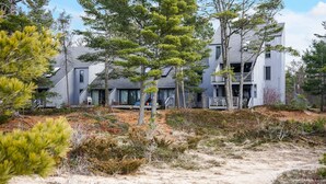 A modern two-story house with a balcony, surrounded by trees and sandy terrain.
