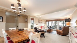 A spacious living room with a wooden dining table, white chairs, a leather sofa, and a television.