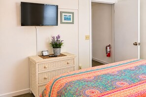 A room with a television mounted on the wall, a bedside table with a clock and a plant, a bed with a colorful bedspread, and a fire extinguisher on the wall. - Key West Oasis on Duval Street : Private Hot Tub • Pool • Parking • 2 Balconies (Key West)