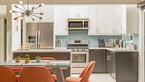 A modern kitchen with white cabinets, a stainless steel refrigerator, a microwave, and a stove. There is a dining area with orange chairs and a glass table.
