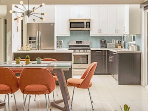 Une cuisine moderne avec des meubles blancs, un réfrigérateur en acier inoxydable, un four à micro-ondes et une cuisinière. On y trouve également un coin repas avec des chaises orange et une table en verre.