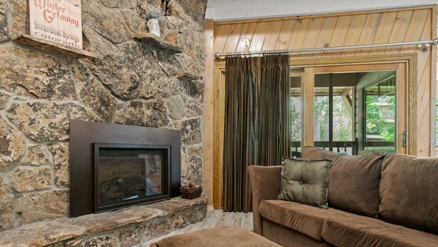 Une cheminée en pierre, un plafond en bois, un canapé marron et une fenêtre avec des rideaux.