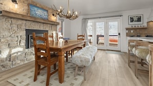 Eine moderne Küche mit Kamin aus Stein, einem Holzespeisetisch und Stühlen. Eine Bank mit Felldecke lädt zum Verweilen ein, darüber hängt eine Chandelier.